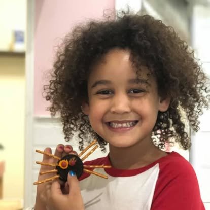 A young girl with curly hair smiling and holding a craft project that resembles a spider made from a cookie, pretzel sticks, and candy.