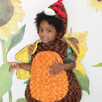 Young girl dressed in a turkey costume, standing in front of a sunflower background, smiling and pointing.