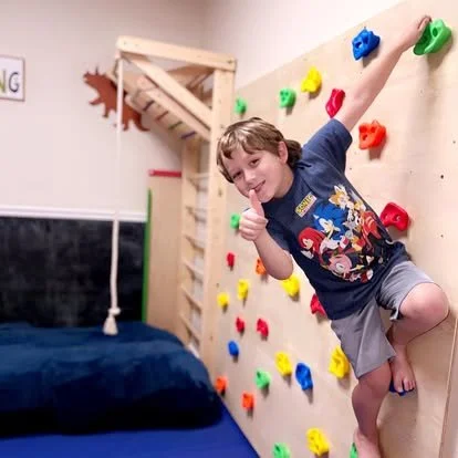 Child smiling and posing on a colorful indoor climbing wall in a bedroom.