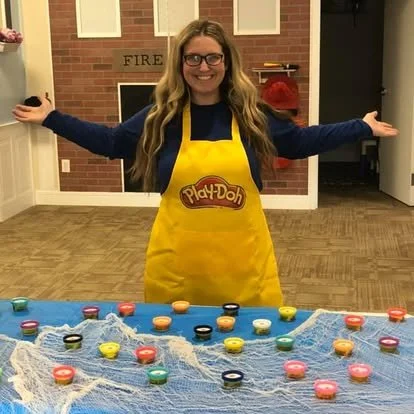 Woman wearing glasses and a yellow Play-Doh apron standing with arms outstretched in a room with a brick fireplace and colorful Play-Doh tubs on a table covered with a blue cloth.