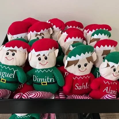 A collection of plush Christmas elf dolls wearing red and green hats, with names like Miguel, Dimitri, Sahle, Alaska, and Oliver embroidered on their outfits, arranged on a shelf.
