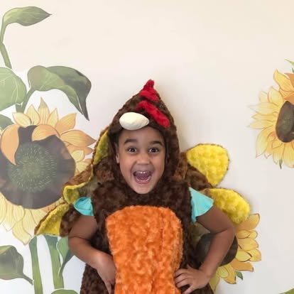 A young girl in a bee costume smiling and posing with her hands on her hips, standing in front of a sunflower mural.