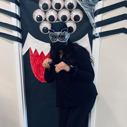 Person wearing a black mask with a panther face design, standing in front of a door decorated with multiple eyeballs and spider web, dressed in black clothing.