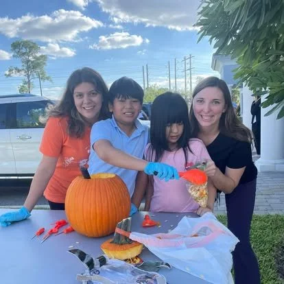 Four children, two girls and two boys, standing around a table outdoors during daytime. They are decorating a carved pumpkin with orange and yellow colors. The children are wearing casual clothes, with one girl in an orange shirt, a boy in a blue shirt, a girl in a pink shirt, and another girl in a black shirt. There are utensils and pumpkin carving tools on the table, and a pumpkin seed bag being used.