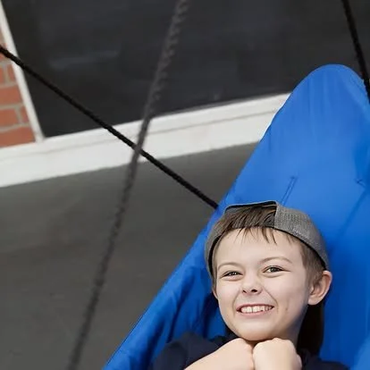 Smiling young boy wearing a gray cap, sitting in a blue swing, outdoors near a building.