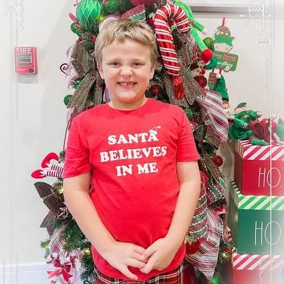 A young boy smiling with hands clasped in front of him, standing in front of a decorated Christmas tree with ornaments, ribbons, and candy canes.