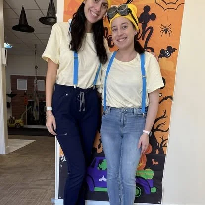 Two women dressed as minions posing together indoors, smiling at the camera, with a Halloween-themed background.