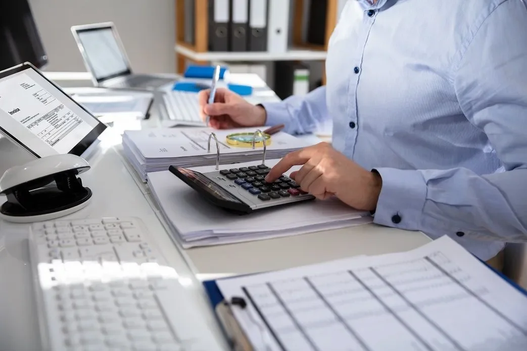 Person using a calculator and writing on documents at a desk with office supplies.
