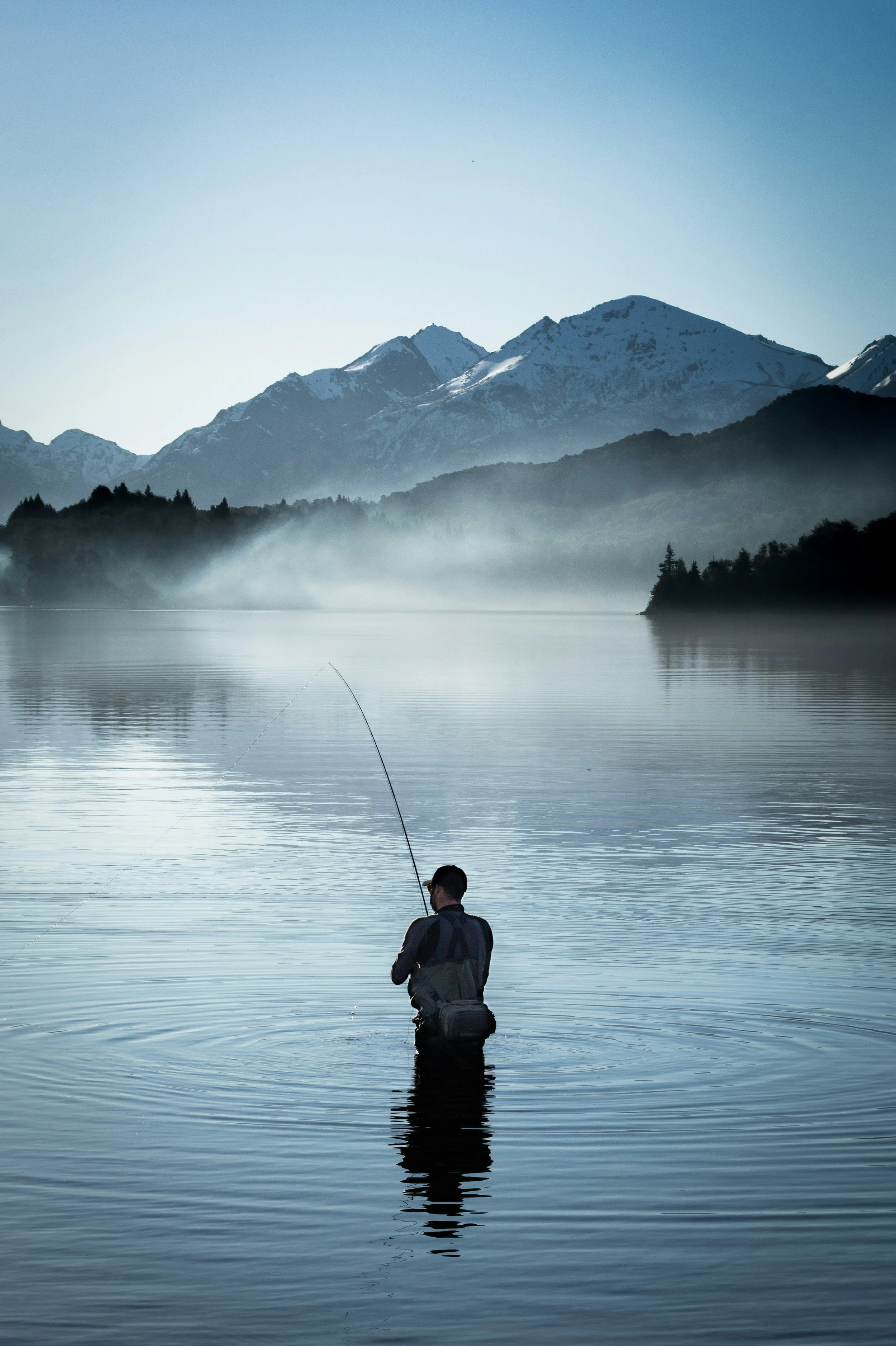 A man stands in a calm lake, fishing with a rod, with a backdrop of snow-capped mountains and mist over the water.