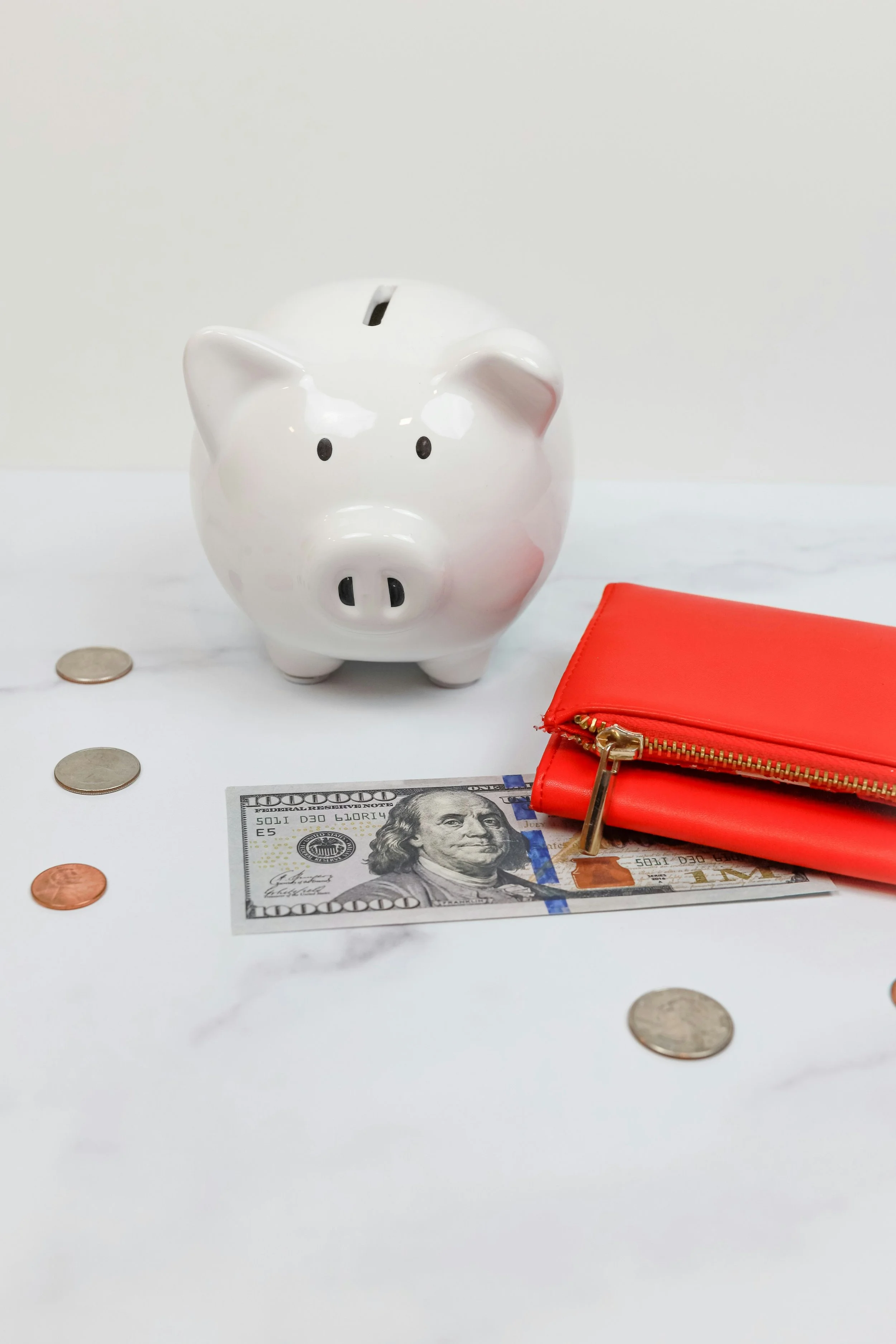 White piggy bank with a coin slot, surrounded by coins, a hundred-dollar bill, and a red wallet on a white surface.