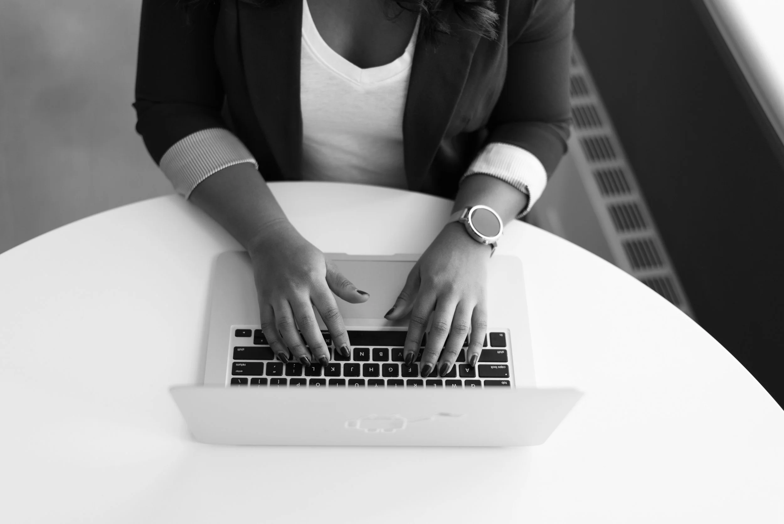 A woman working on a laptop at a white table, wearing a watch and a blazer.
