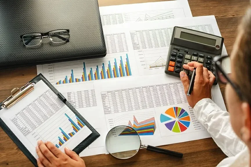 Person analyzing financial reports, charts, and graphs with a calculator, magnifying glass, and clipboard on a wooden desk.