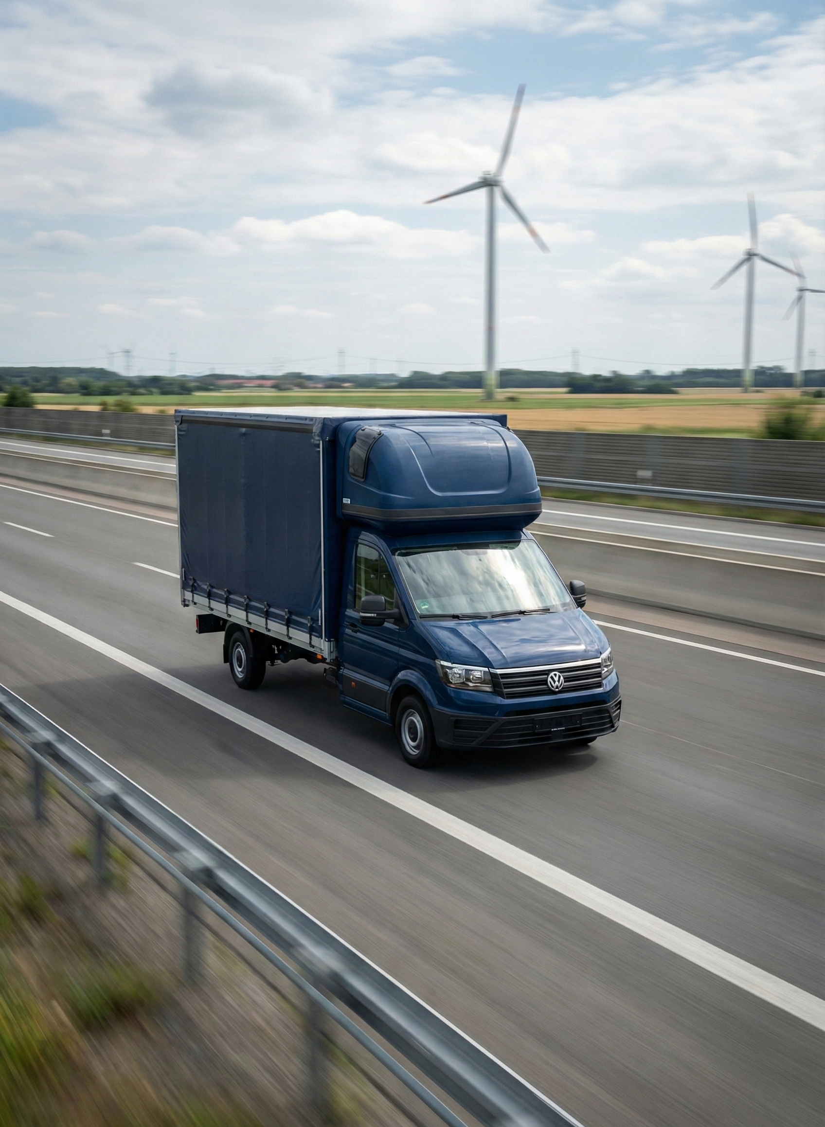 Ein blauer Lieferwagen fährt auf einer Autobahn mit Windkraftanlagen im Hintergrund.