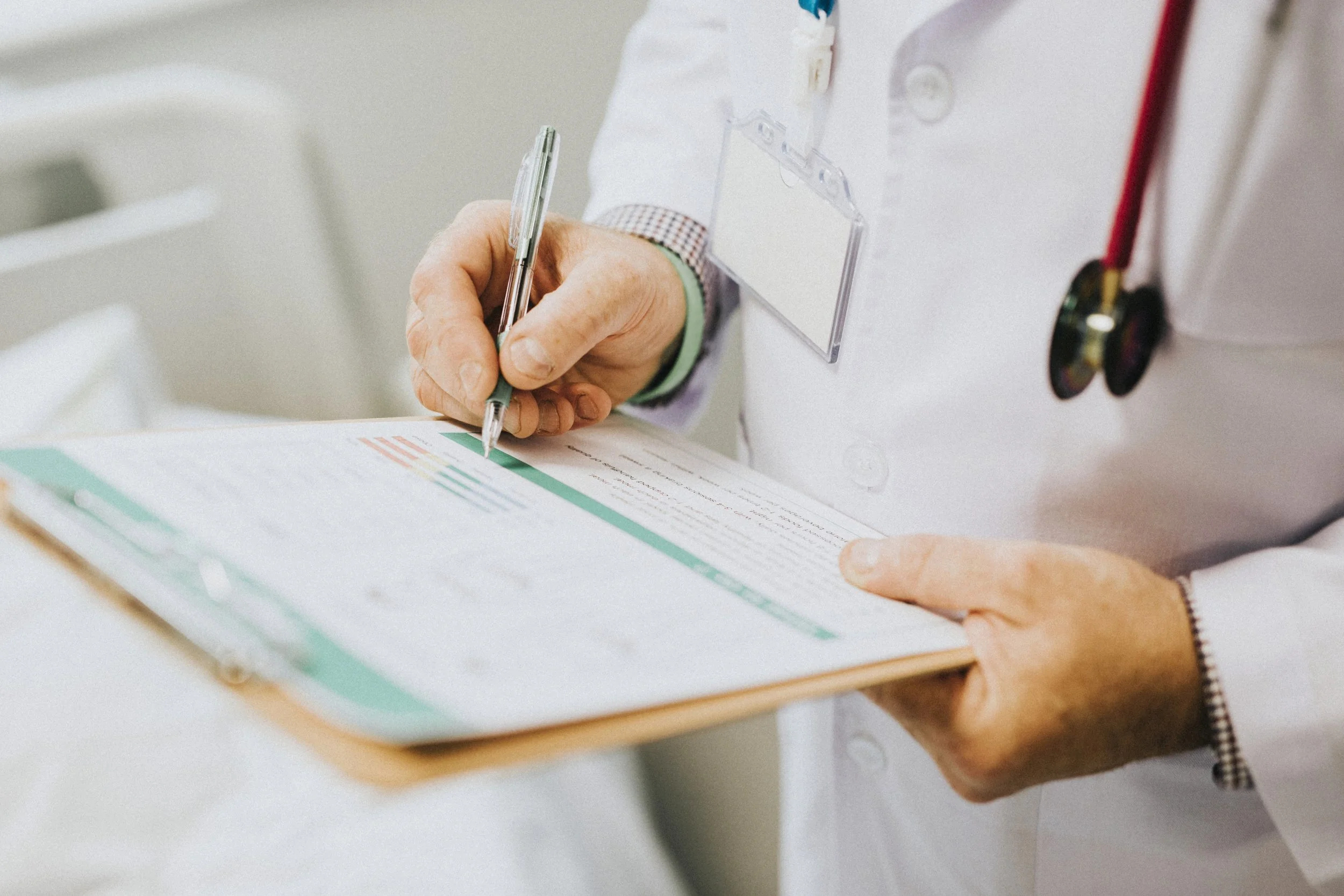 A healthcare professional in a white coat is holding a clipboard and writing on a document with a pen. The person is wearing a stethoscope around their neck.