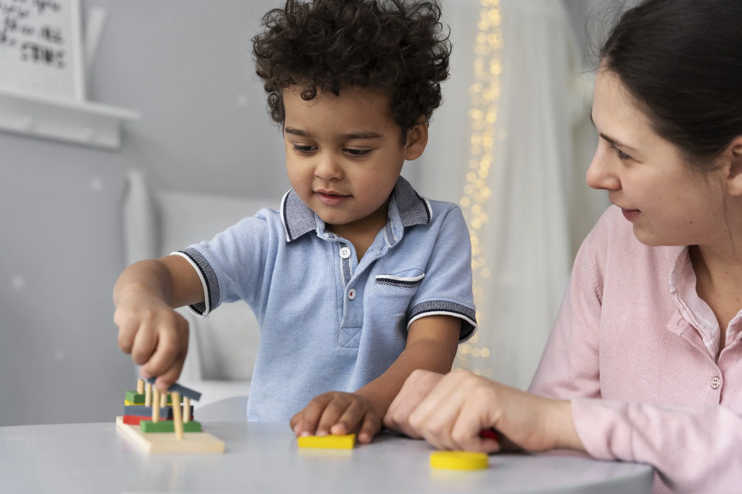 A young boy playing a board game with an adult woman, both focused on the game pieces on a table in a brightly lit room.