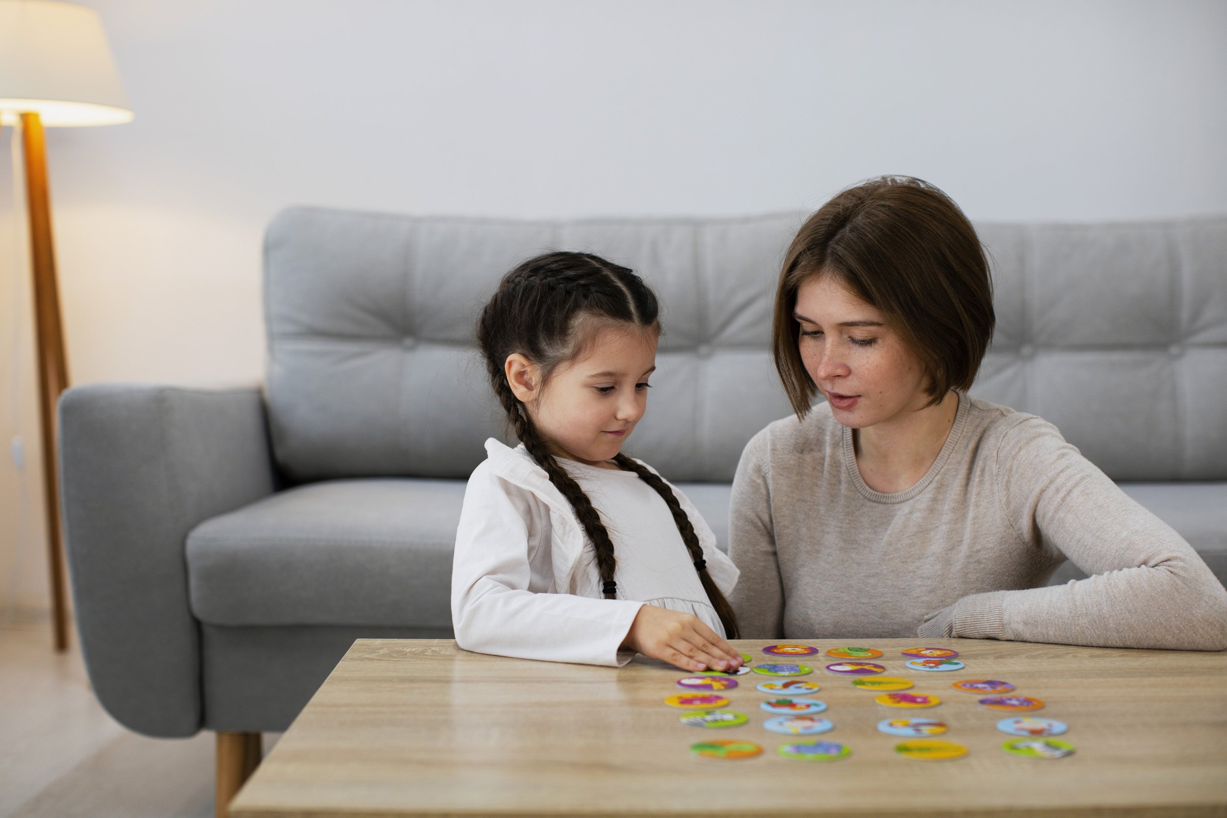 A woman and a young girl playing a colorful alphabet matching game on a wooden table in a living room. The woman has brown hair and wears a light gray shirt. The girl has dark braided hair and wears a white shirt. A gray sofa and a white lamp are in the background.