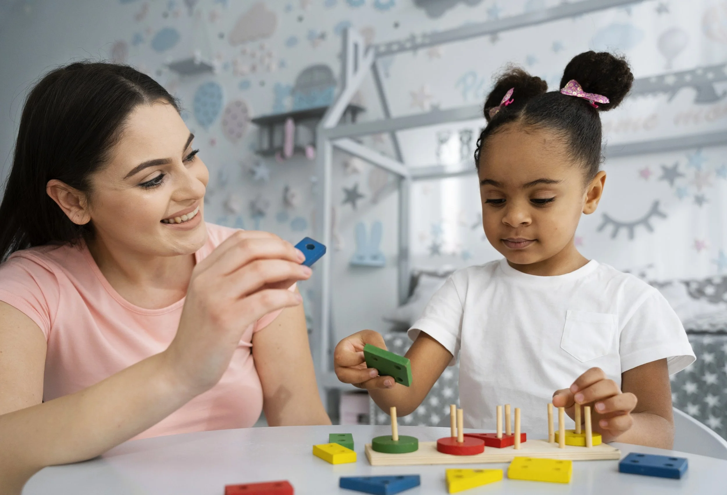 A woman and a young girl playing a tabletop educational game with colored blocks and pegs in a child's bedroom with a star and moon themed wallpaper.