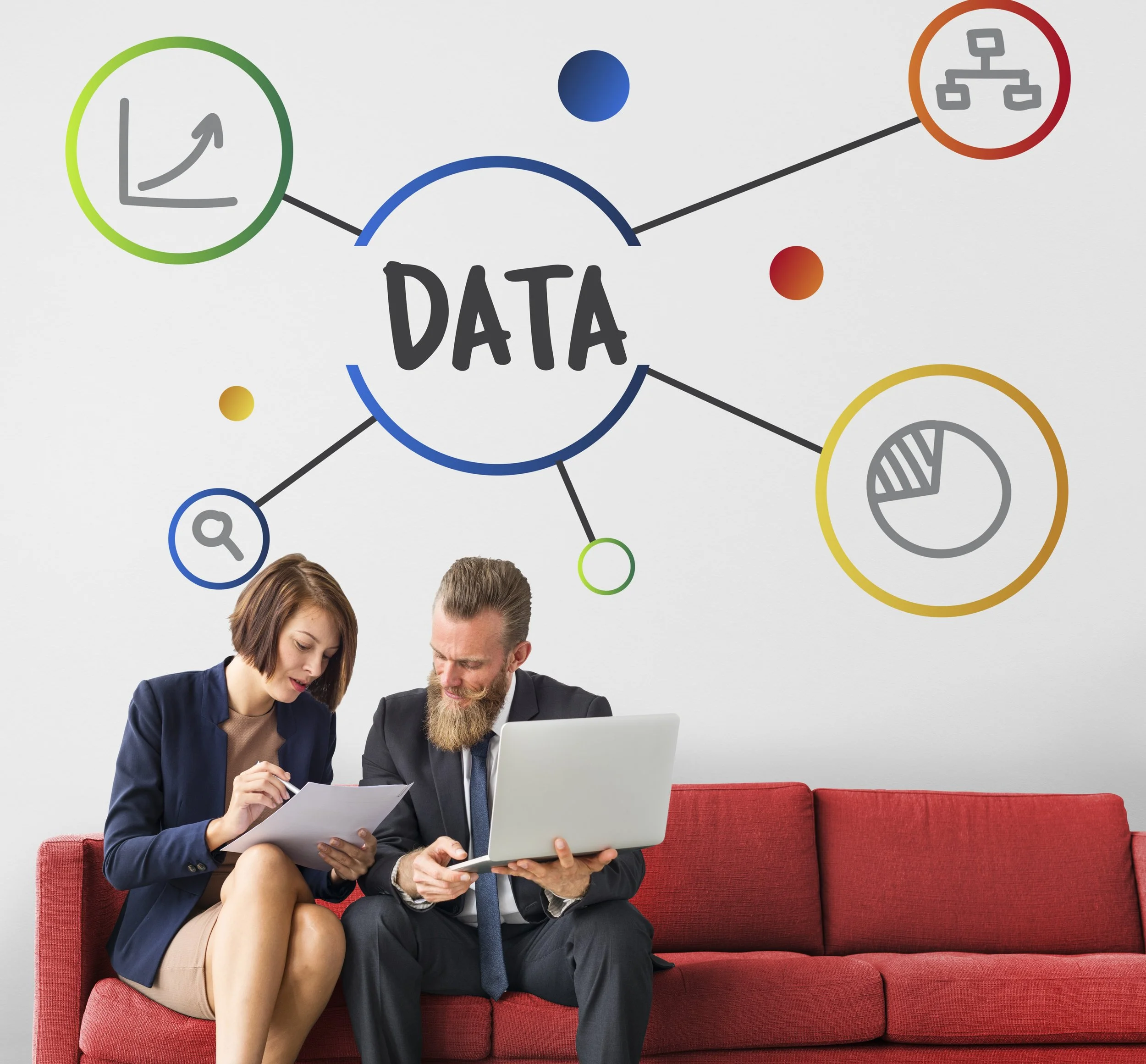 Two professionals, a woman and a man, sitting on a red sofa examining documents and a laptop, with a graphic of connected data icons and charts on the white wall behind them.