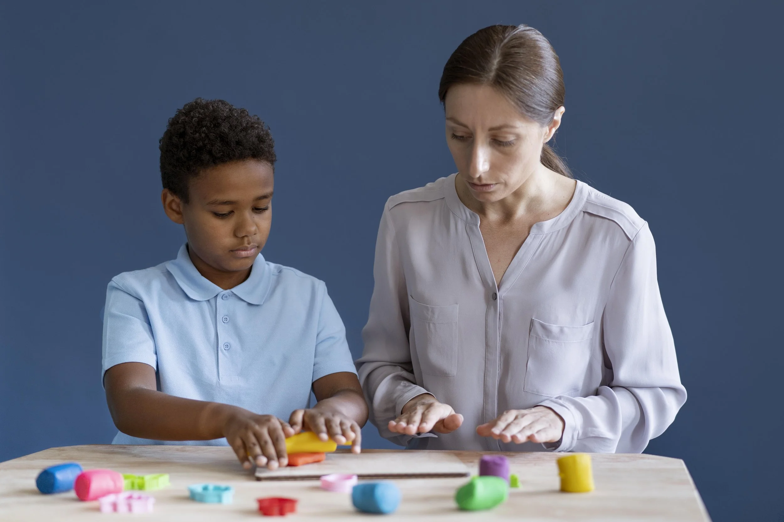 A young boy and an adult woman playing with colorful modeling clay at a wooden table against a blue background.