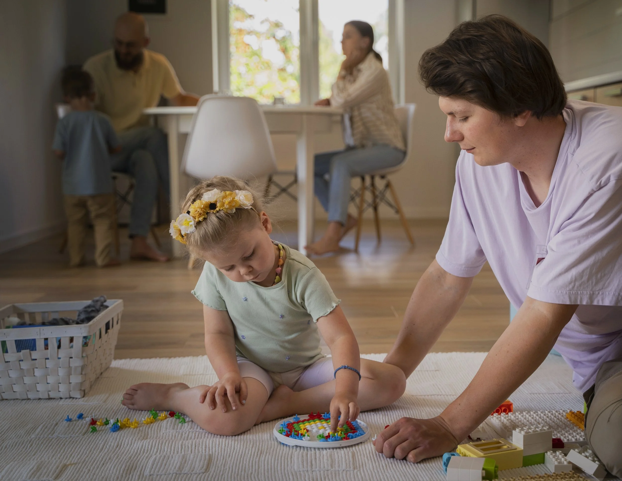 A young girl with a flower headband sitting on a rug, playing with colorful building blocks, while an adult kneels beside her, helping, in a cozy home setting.