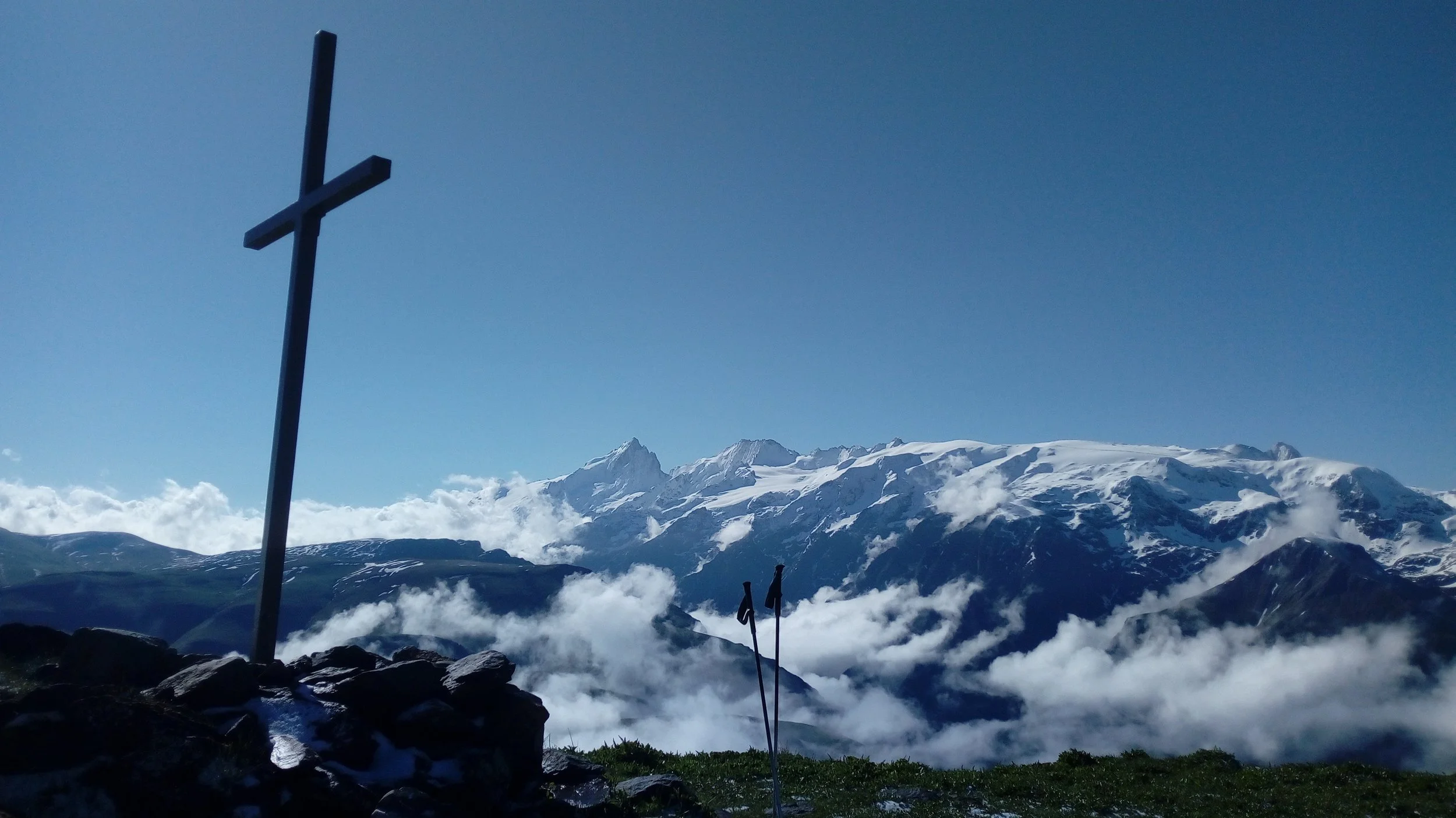 Une croix située sur une montagne, avec des montagnes enneigées en arrière-plan et un ciel clair.