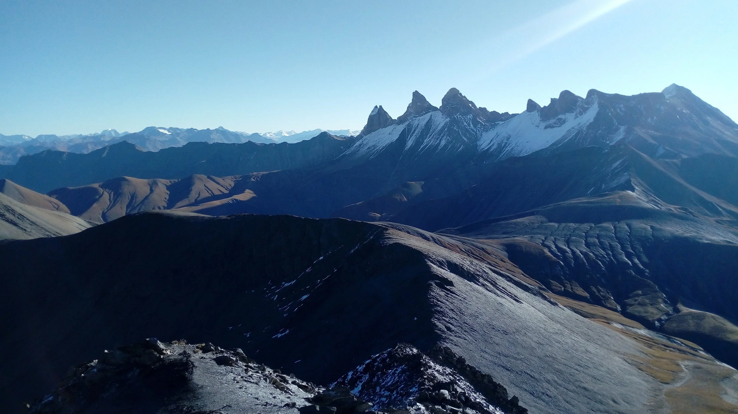 Vue panoramique de montagnes enneigées avec sommets rocheux et vallées colorées, sous un ciel clair.