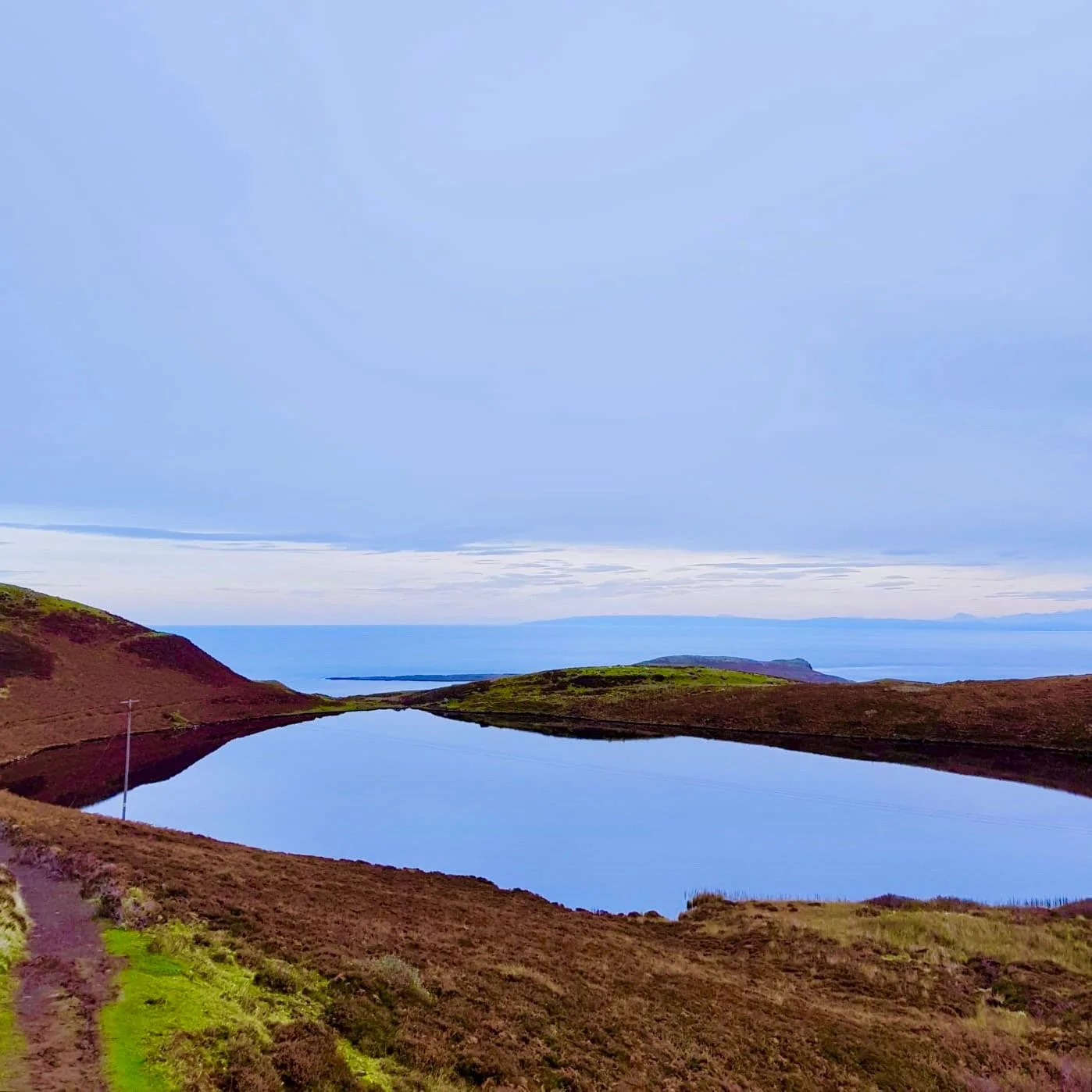 Paysage côtier avec un lac au premier plan, des collines verdoyantes et une mer calme en arrière-plan, sous un ciel nuageux.