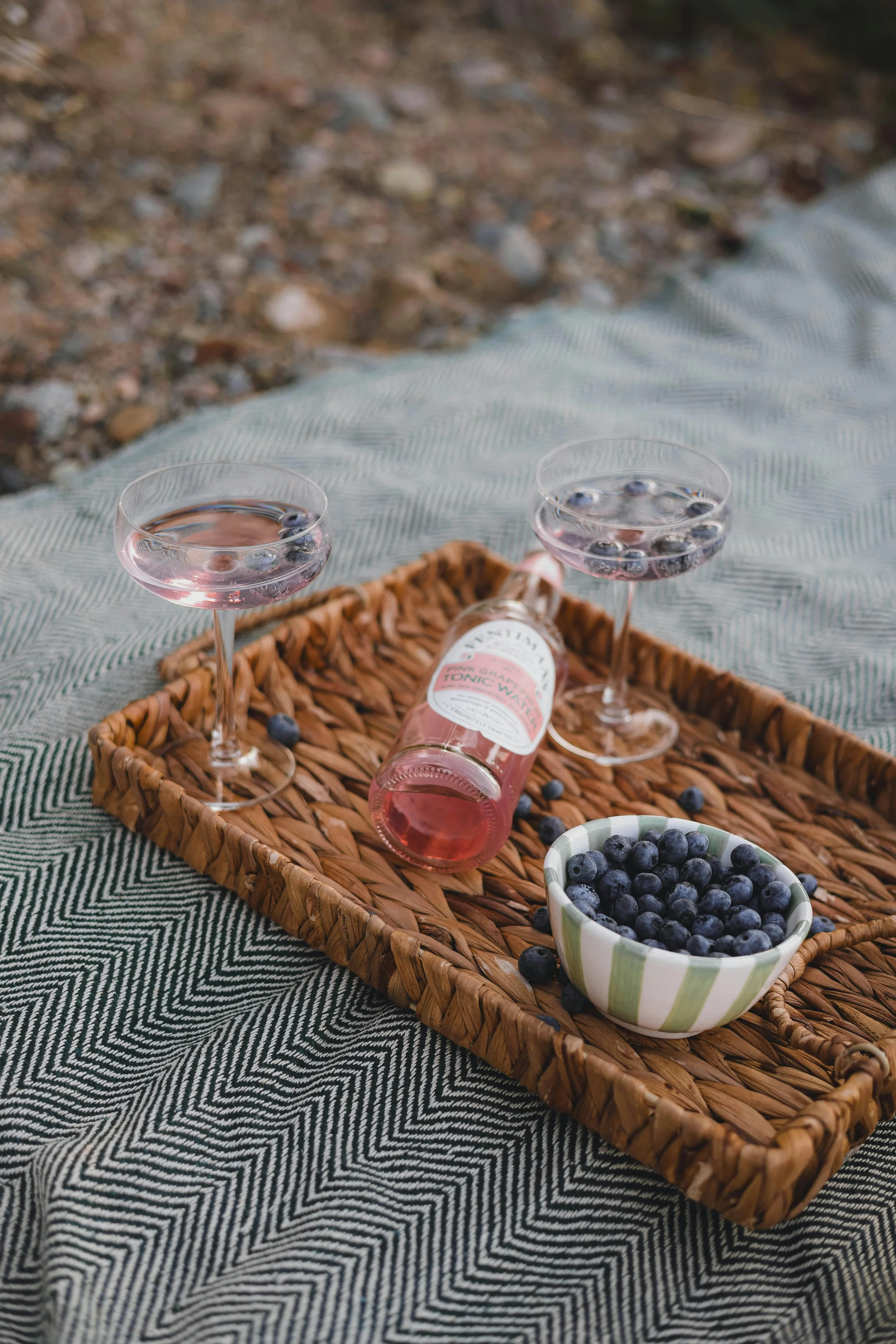 A wicker tray holding a pink tonic water bottle and two glasses with pink drinks, surrounded by blueberries, on a picnic blanket outdoors.