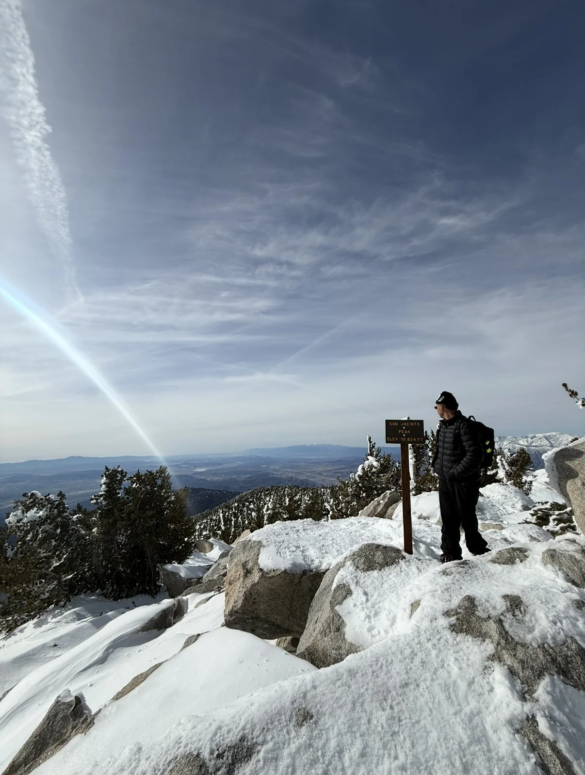 A person standing on snow-covered rocks beside a sign at the peak of San Jacinto, with a mountain range in the distance and a partly cloudy sky.