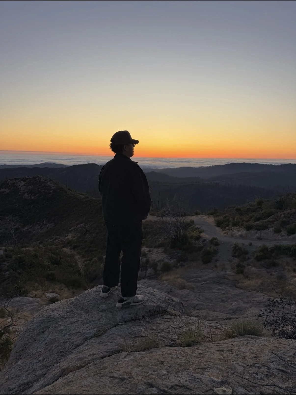 A person standing on a rocky outcrop, overlooking a mountain landscape at sunset, with a vibrant orange and yellow sky and clouds in the distance.