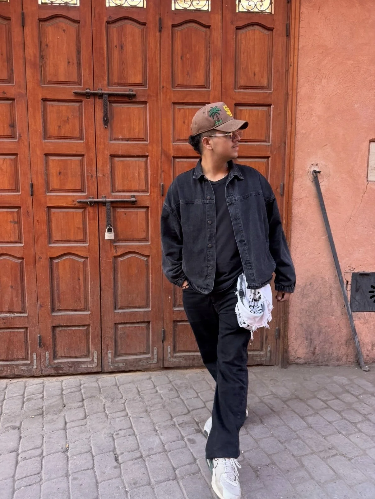 A person wearing a brown cap with palm tree and 'S' patches, an oversized black denim jacket, black pants, white sneakers, and glasses, standing in front of a wooden door with a padlock, on a cobblestone street.