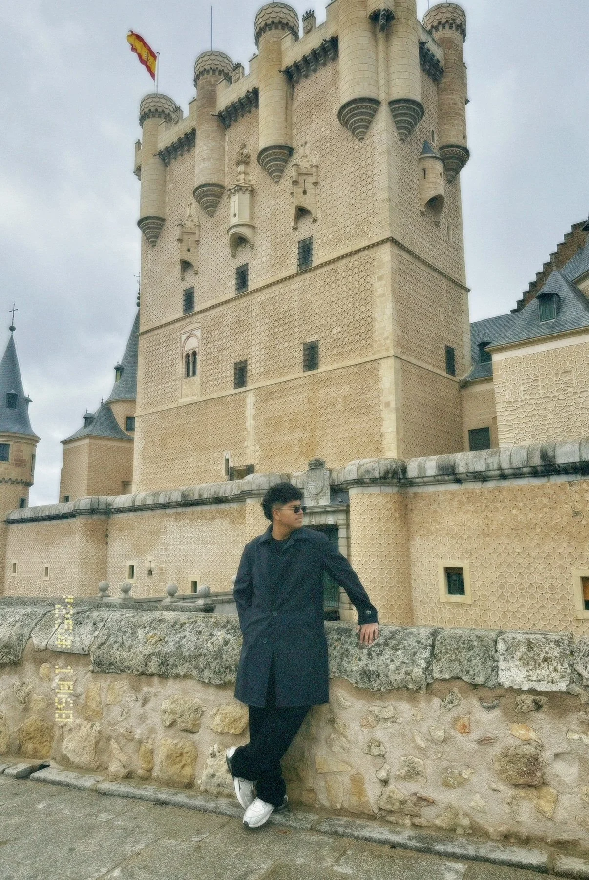 A person wearing a black coat, black pants, white sneakers, and sunglasses leaning against a stone wall in front of a castle with beige, textured walls, turrets, and a castle flag.
