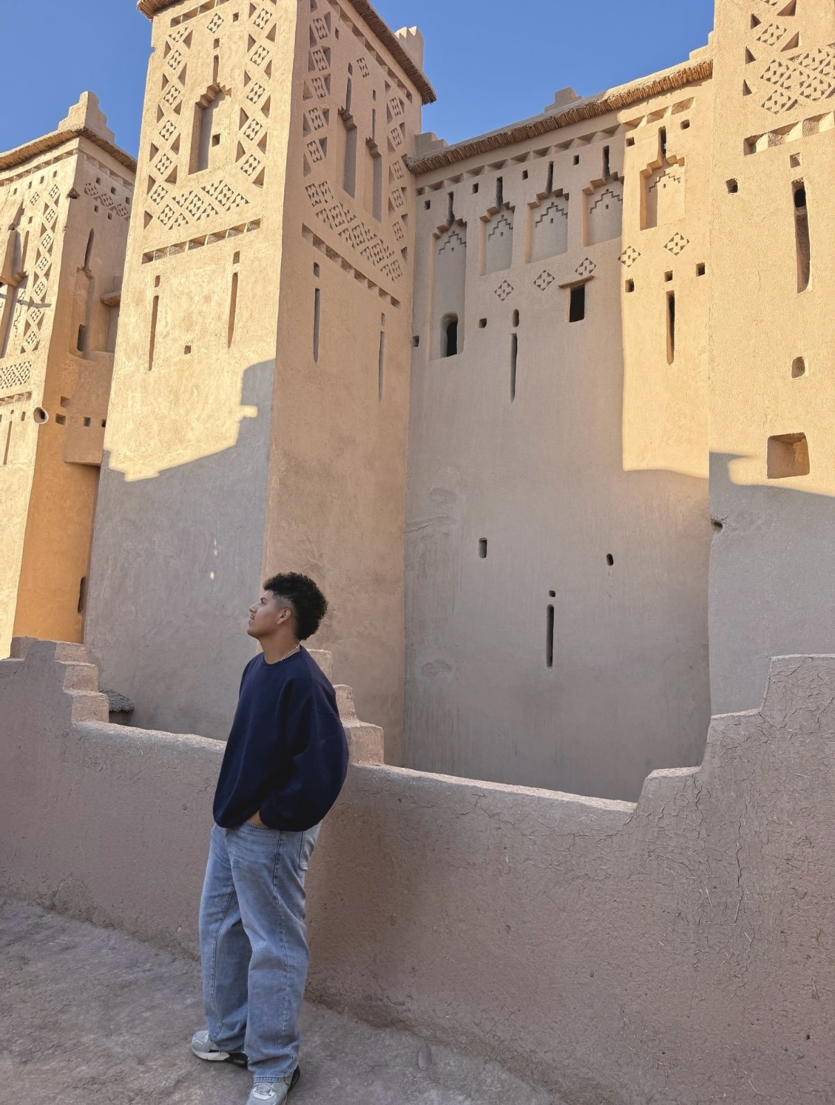 A young man wearing a navy blue sweatshirt and light jeans stands with hands in pockets, looking up at a traditional Moroccan kasbah with beige, intricately decorated walls and small windows, under a clear blue sky.