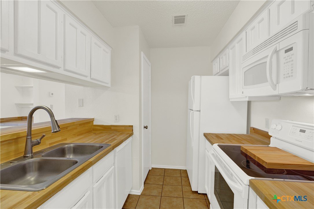 Kitchen with white cabinets, wooden countertops, white appliances including microwave and refrigerator, sink with two basins, electric stove, and tiled floor.