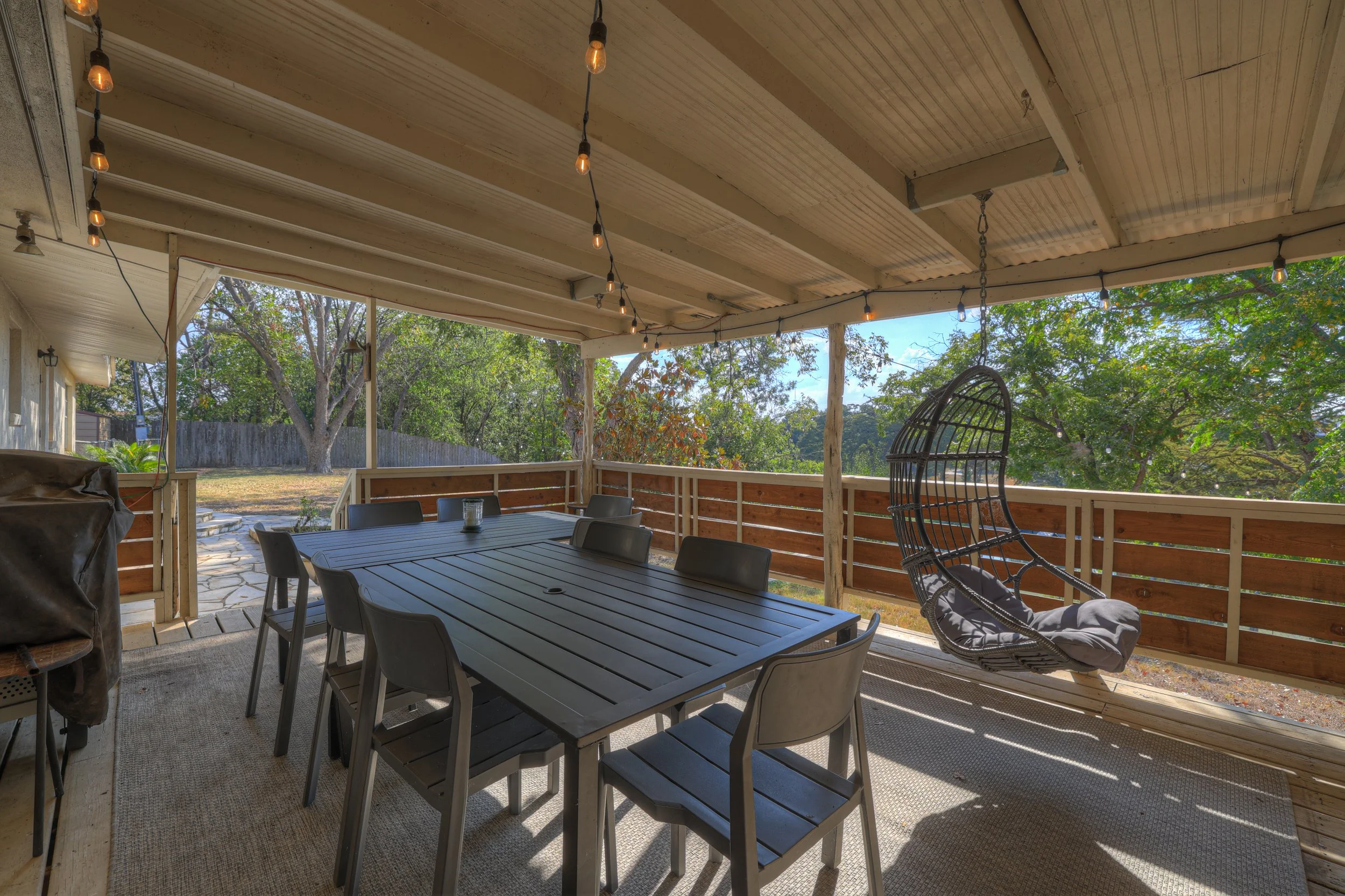 Outdoor porch with wooden ceiling, hanging string lights, dining table with multiple chairs, and a hanging hanging egg chair with a cushion, overlooking a backyard with trees and a stone pathway.