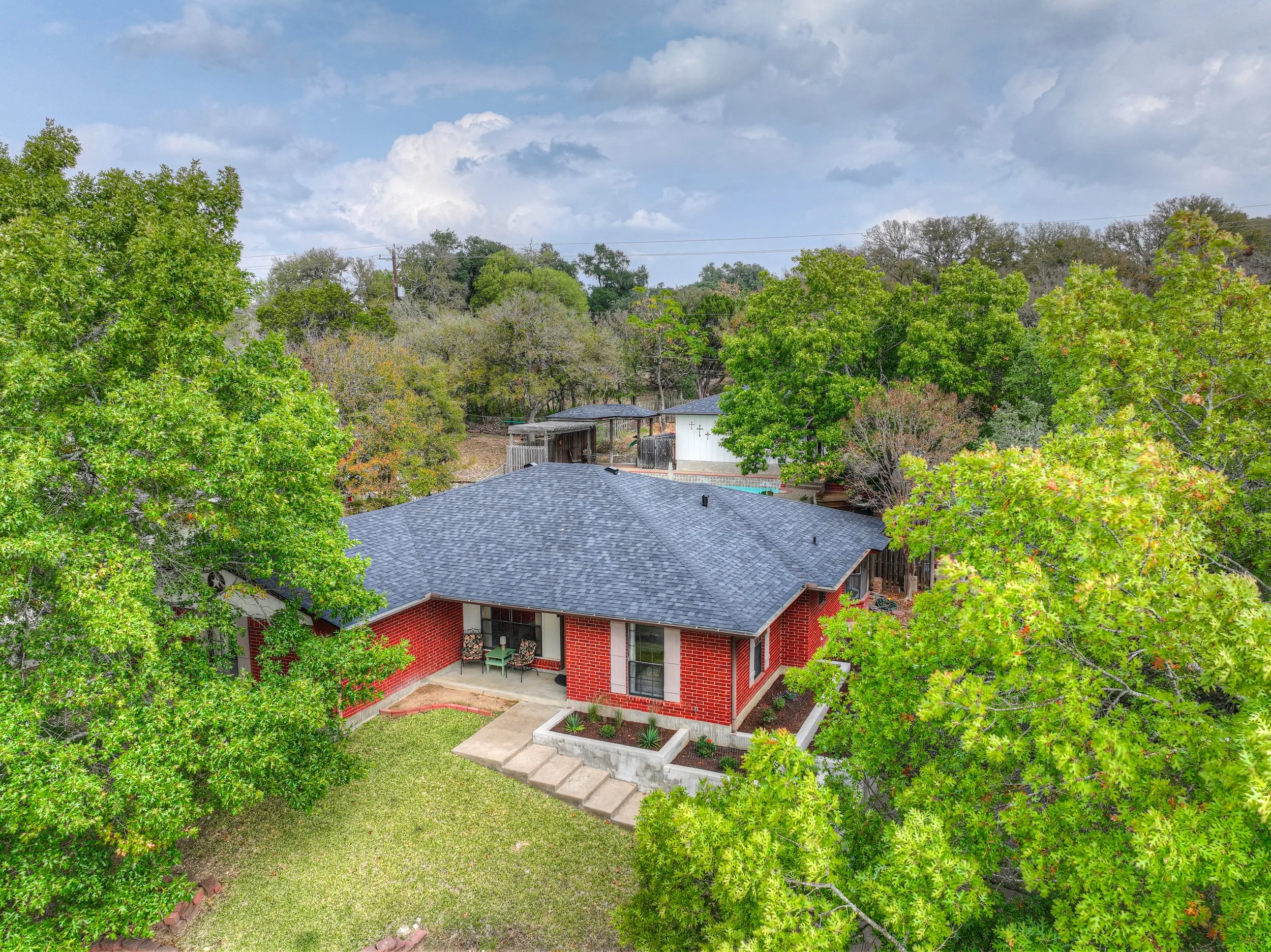 Aerial view of a red brick house with a dark gray shingled roof, surrounded by green trees and a lush backyard with a lawn and a patio with outdoor furniture, under a partly cloudy sky.