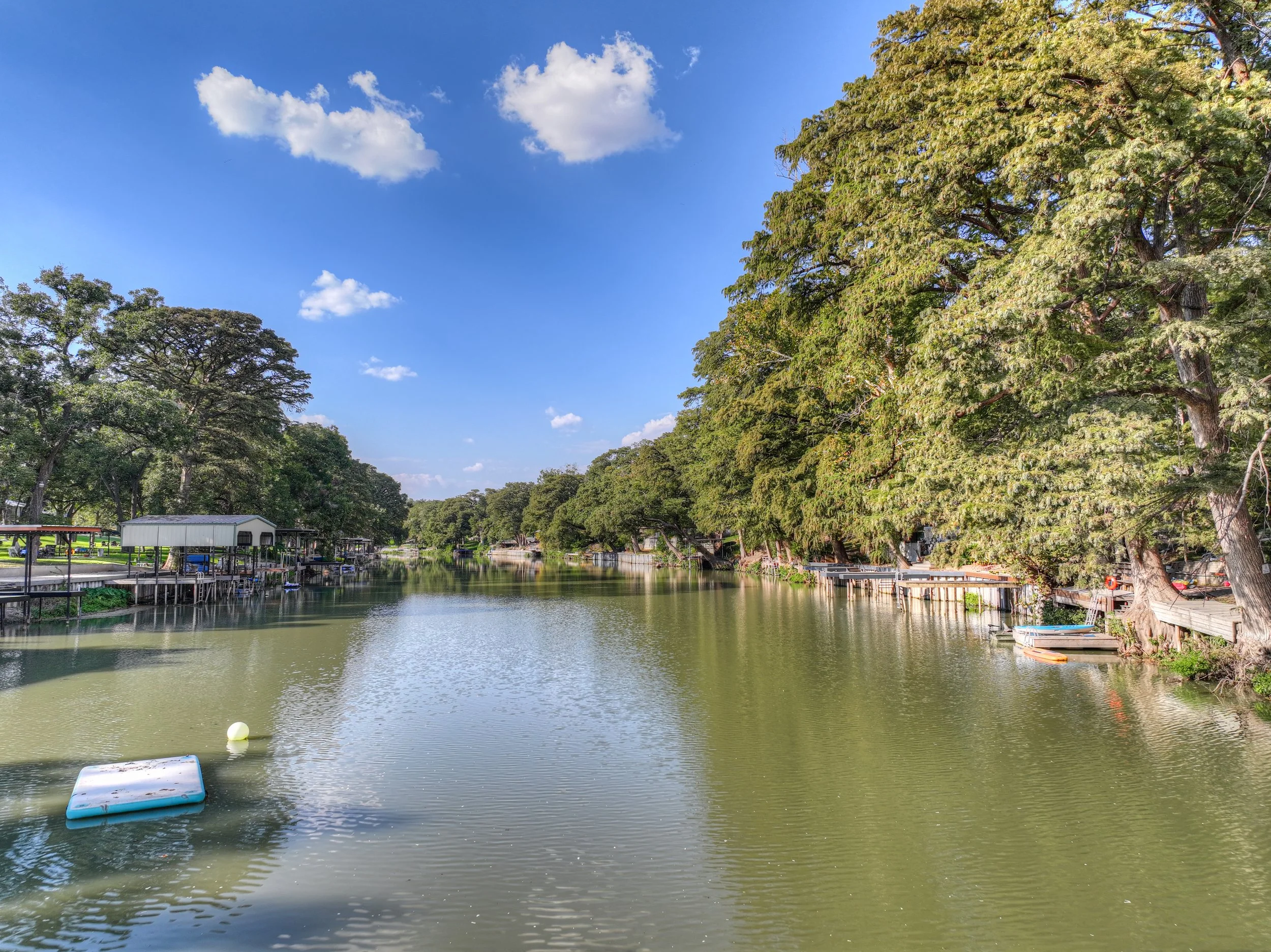 A peaceful river scene with calm water reflecting the blue sky and a few white clouds, surrounded by lush green trees and wooden docks with boats on both sides.