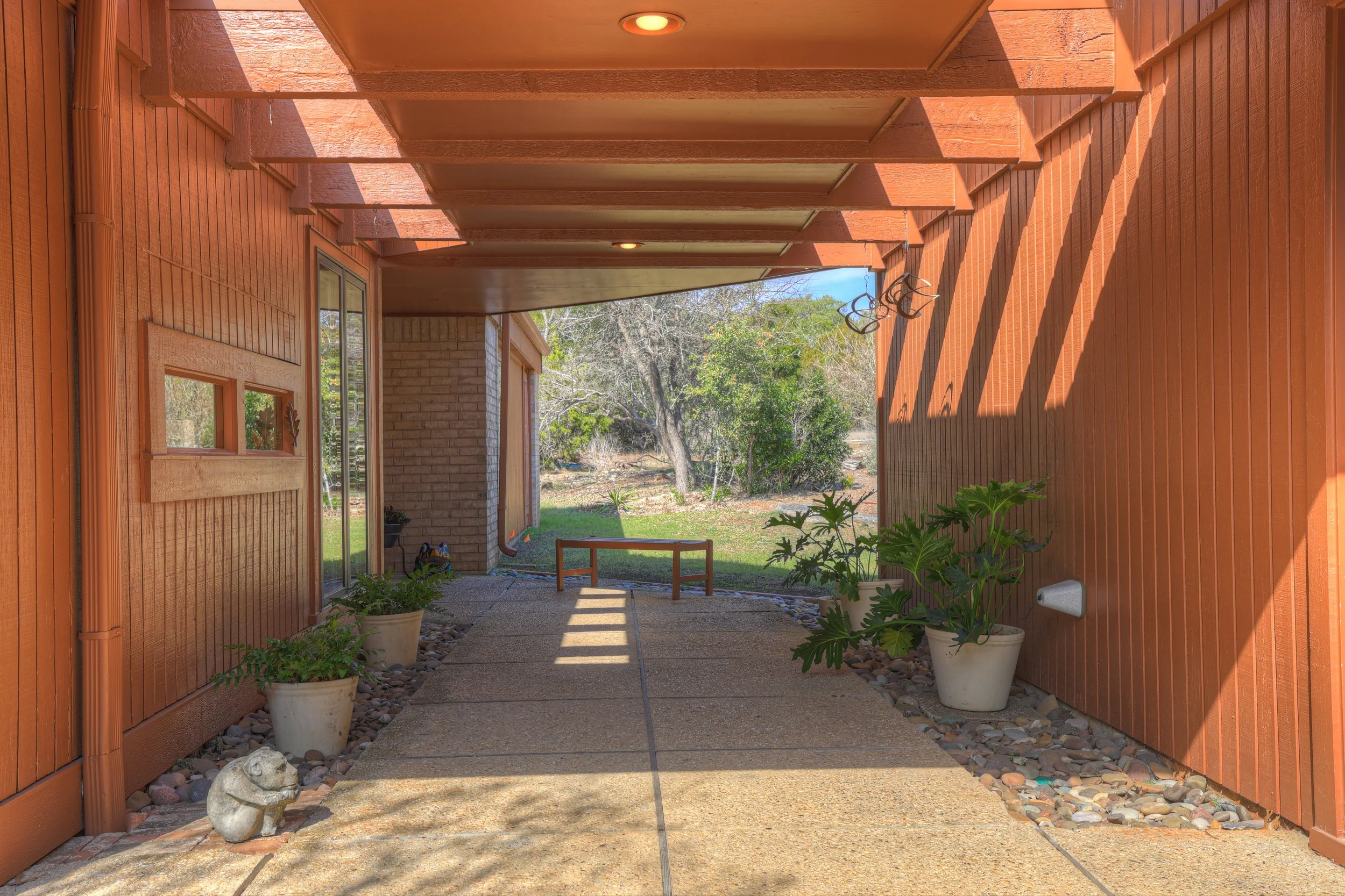 View of a covered porch area with brick and wood siding, potted plants, and a garden beyond, with trees and grassy yard in the background.