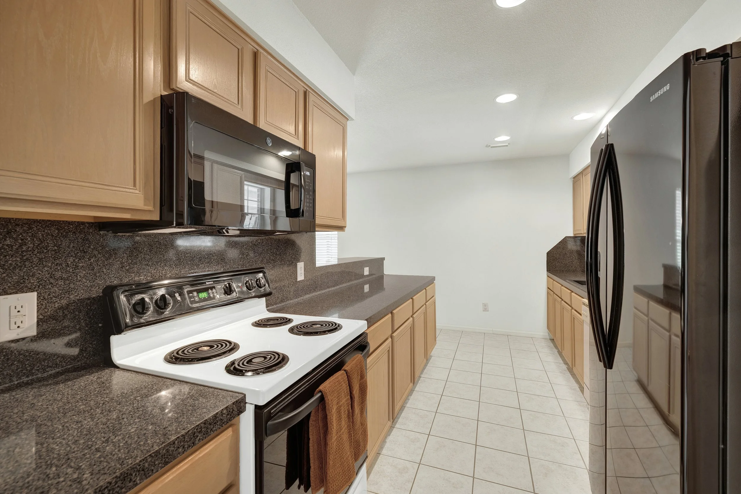 Kitchen with wooden cabinets, black microwave, white stove with coil burners, brown towels hanging on the oven handle, black refrigerator, tiled floor, and white walls.