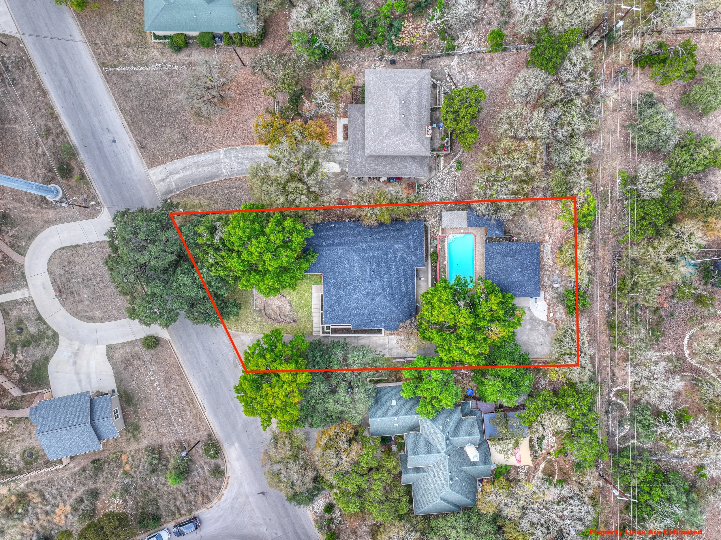 Aerial view of a residential property outlined with a red property line. The property includes a house with a dark roof, a backyard with a swimming pool, and several trees. Surrounding the property are neighboring houses, roads, and utility lines.