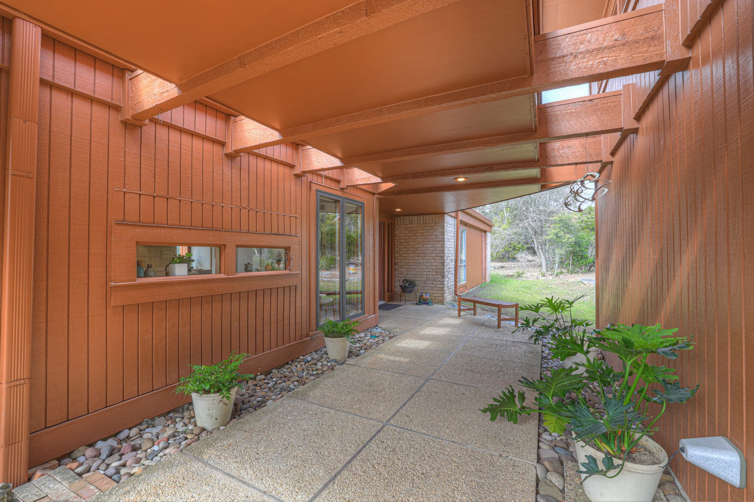 Covered patio area with potted plants, brick and wood exterior, and view of a grassy yard with trees in the background.