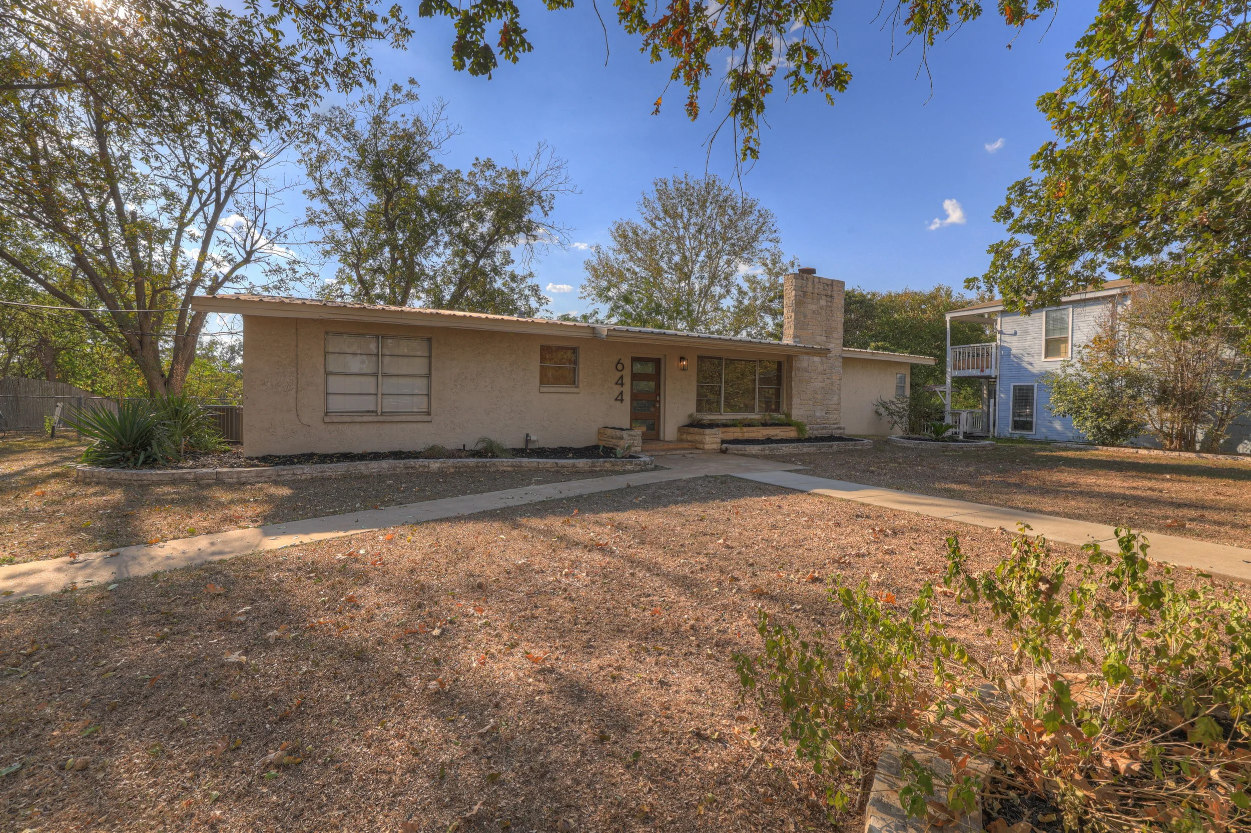 Single-story house with a beige exterior, stone chimney, and a small front porch, surrounded by leafless trees and dry yard, with a neighboring blue house to the right.