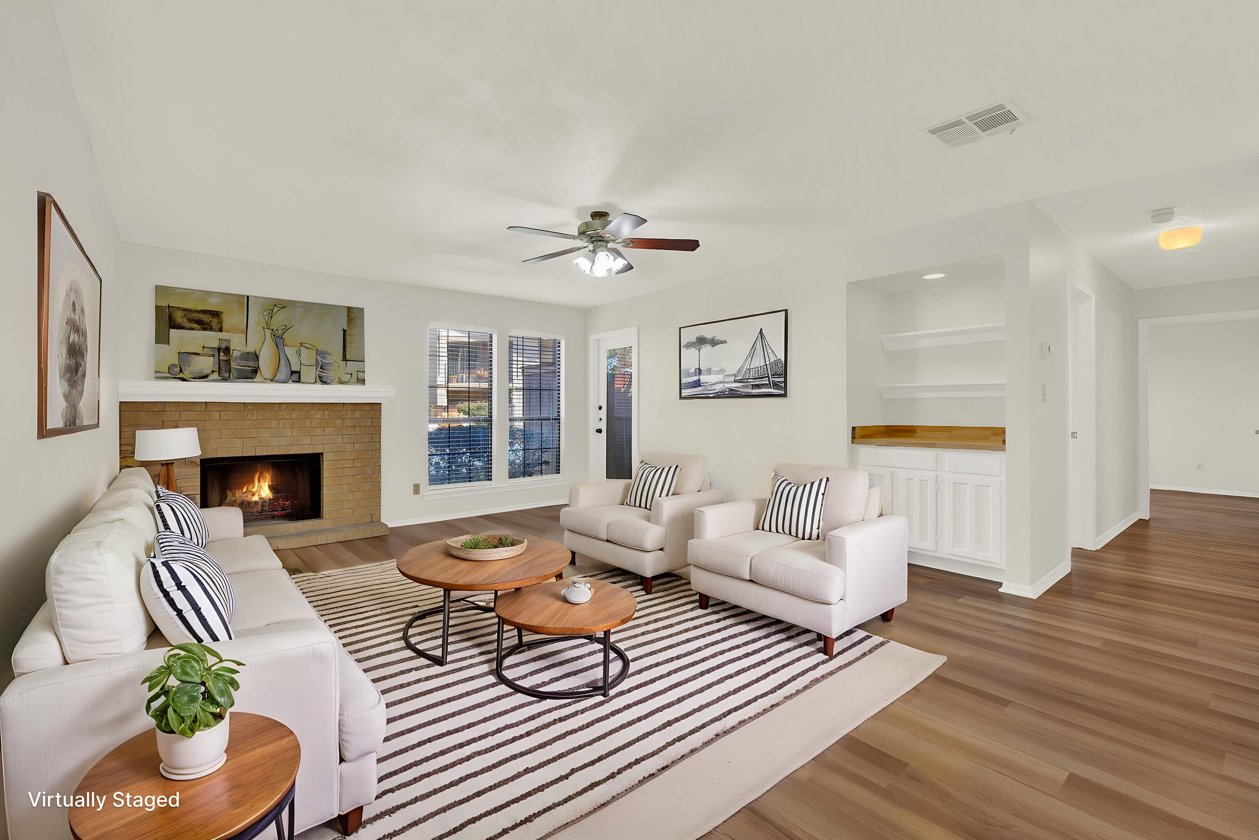 Living room with white sofas, striped pillows, a striped rug, a brick fireplace with a fire, artwork on white walls, wooden flooring, and large windows