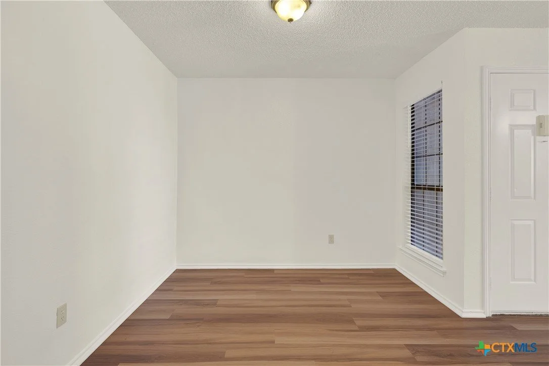 Empty room with white walls, wooden floor, two windows with blinds, ceiling light fixture, and door.