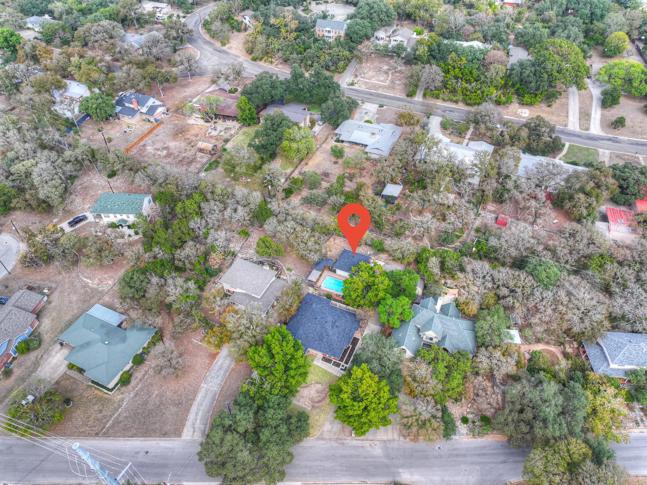 Aerial view of a suburban neighborhood with houses, trees, and streets. A house with a swimming pool is marked with a red location icon.