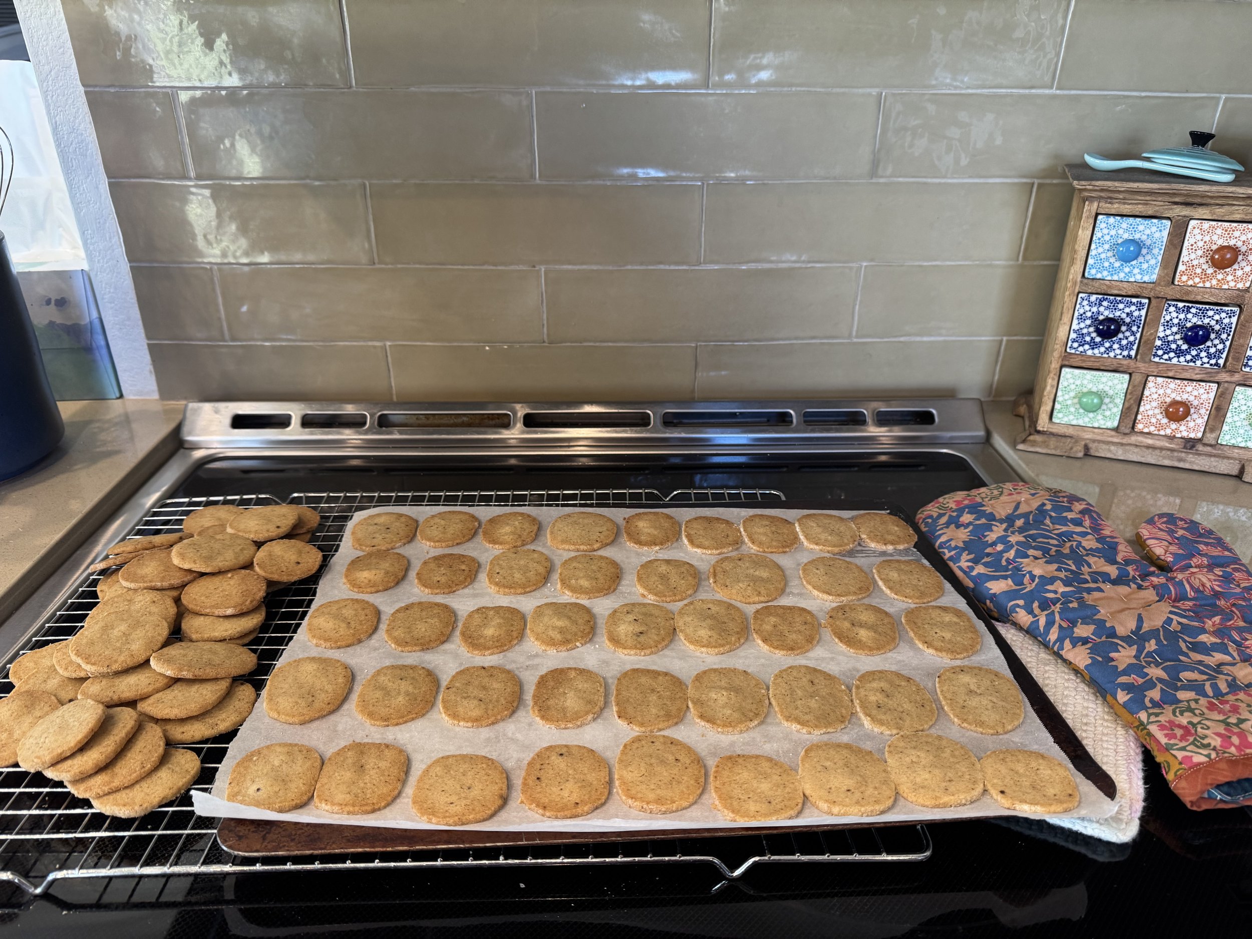 Baked cookies cooling on racks on a kitchen stove, with oven mitts and a colorful storage box on the counter.