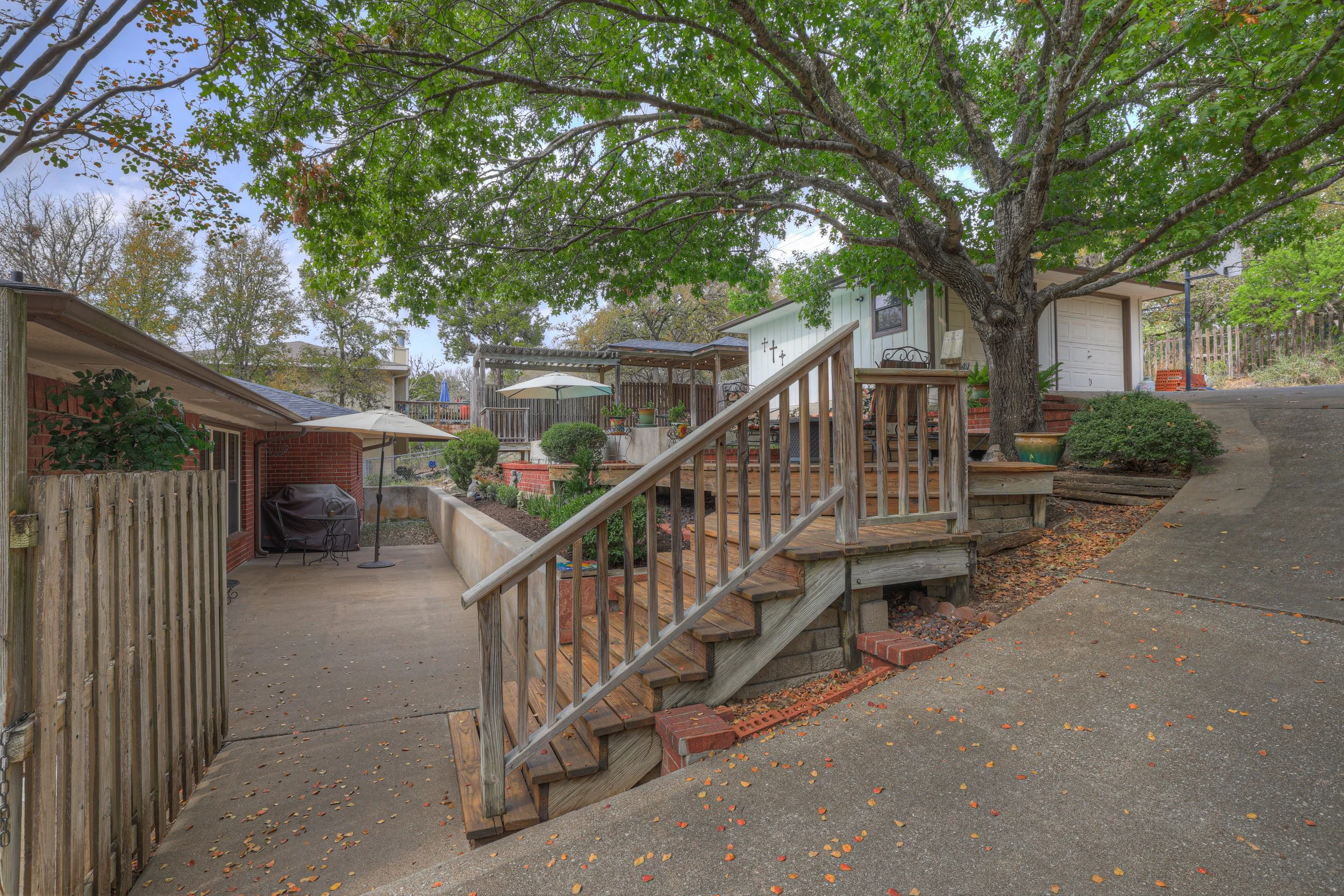 Backyard patio with wooden stairs, large tree, landscaped garden, umbrellas, and a shed in suburban neighborhood.