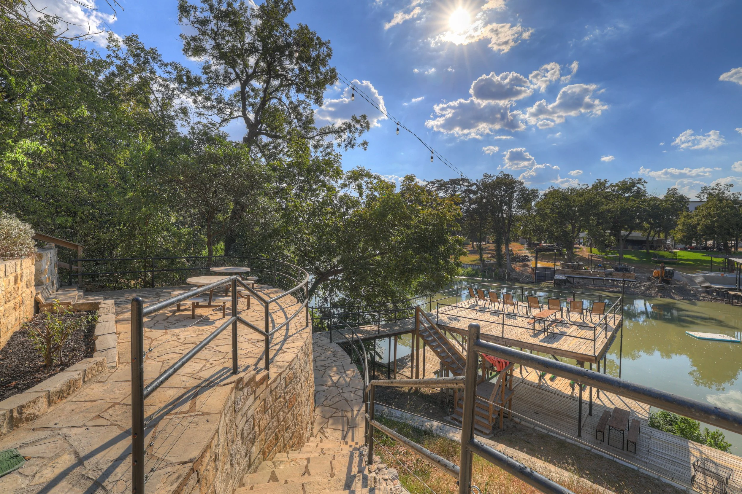 Outdoor patio area with a brick walkway, railing, and a staircase leading down to a lower deck with chairs, overlooking a pond with ducks, trees, and a sunny sky with clouds.