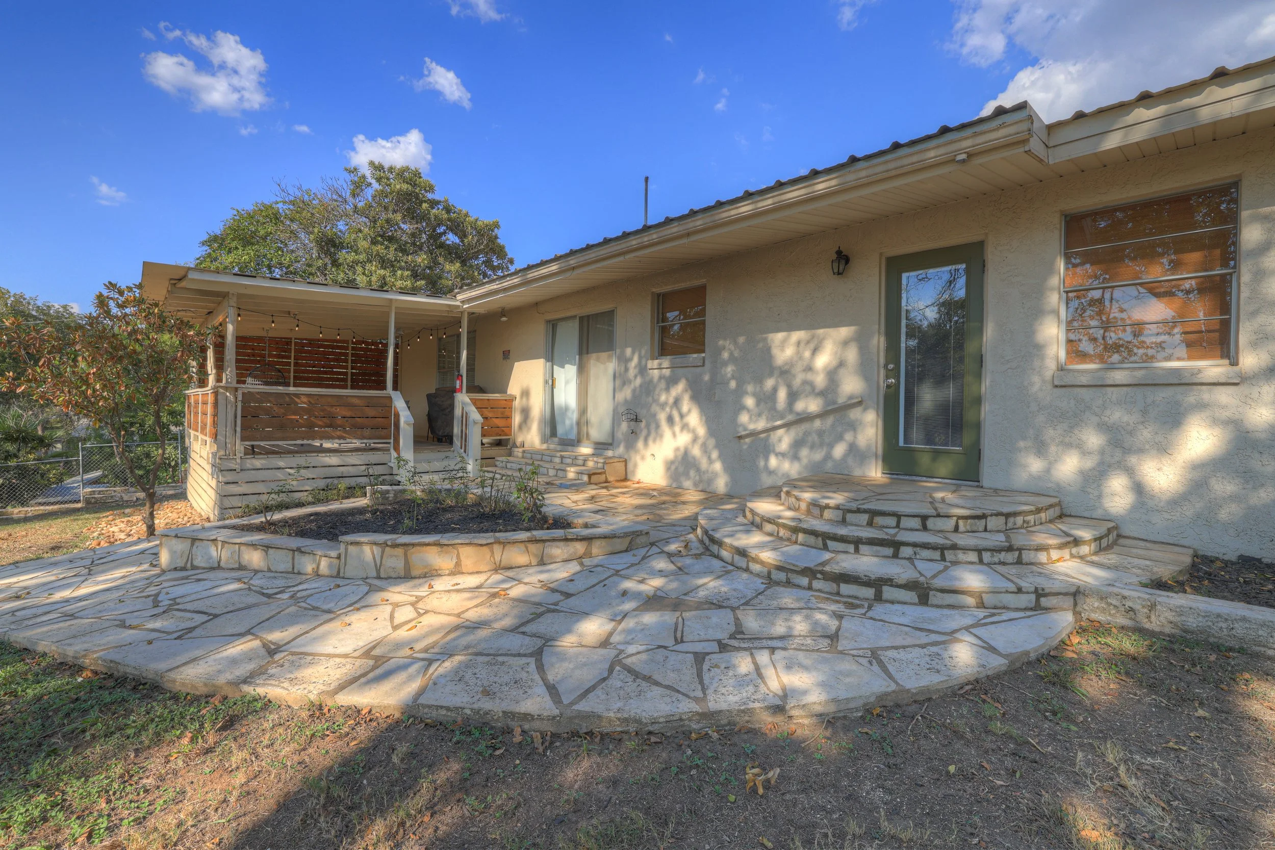 Backyard patio with stone flooring, steps, and a small garden bed with plants, attached to a beige house with green door and windows, and a covered porch area with string lights.
