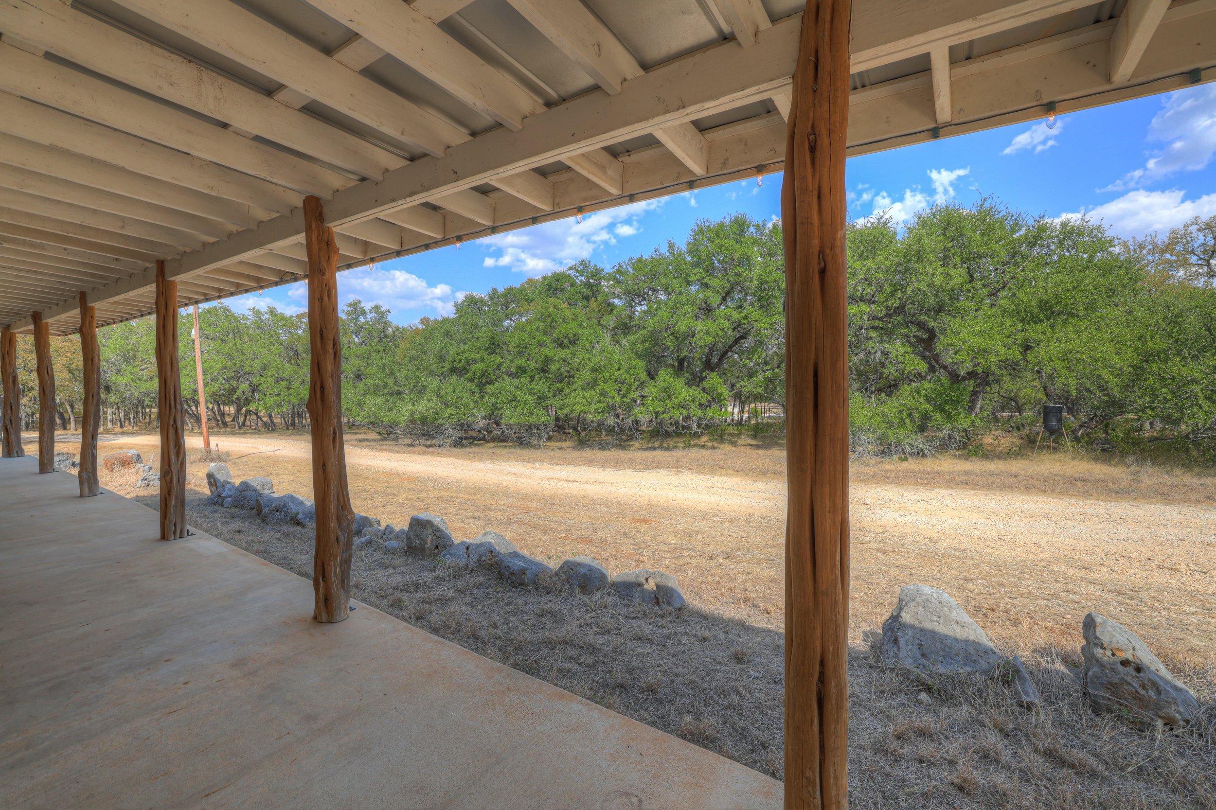 View from a covered porch with wooden posts, overlooking a dirt ground and green trees under a blue sky with scattered clouds.